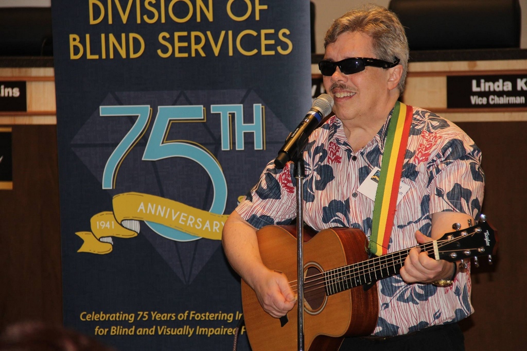 Dave Hillebrandt plays his guitar and sings while standing in front of a DBS 75th anniversary banner 